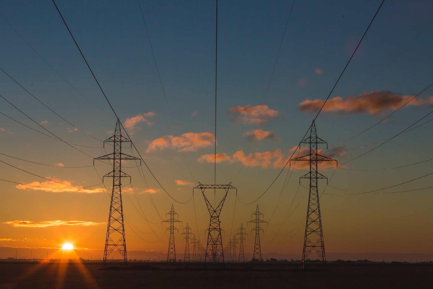 A high-voltage transmission corridor at sunset, with lattice towers receding in a line across flat open terrain and conductors sweeping between them.